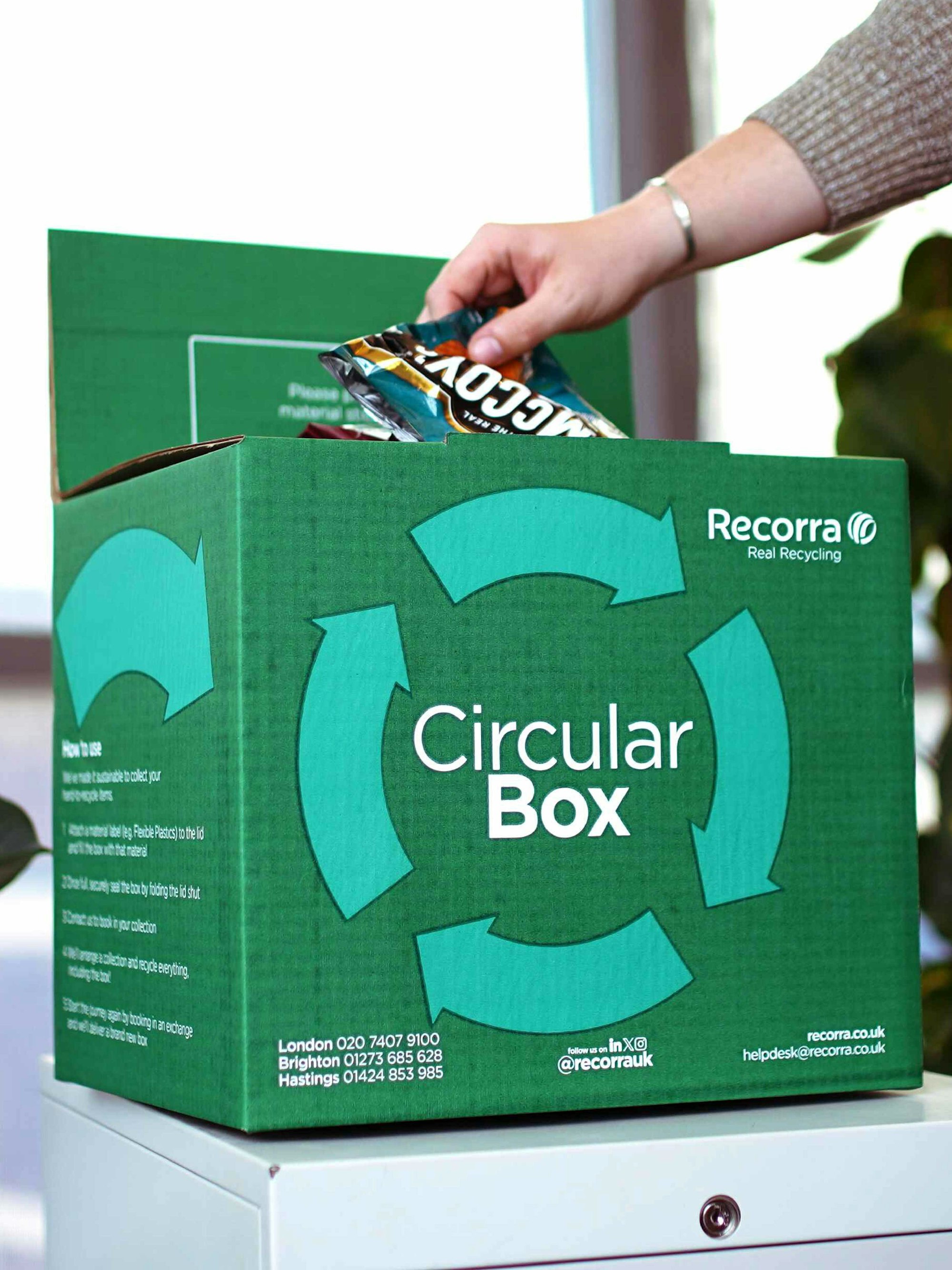 Green circular box for flexible plastics with a hand placing a McCoys crisp packet inside.