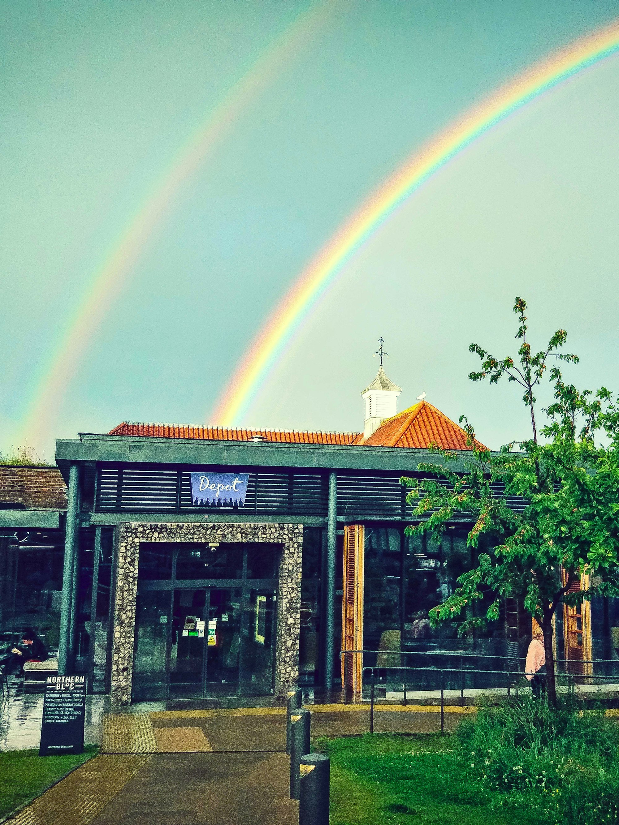 A vibrant double rainbow arcs over a Depot, Lewes their sign reading "Depot." A tree and green grass are in the foreground, adding a fresh atmosphere.